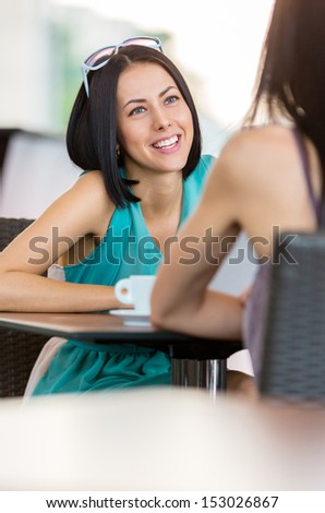 Girl wearing blue dress and sunglasses sits at the table of the bar and talks with friend