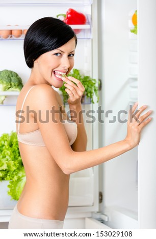 Young woman eating near the opened fridge full of vegetables and fruit. Concept of healthy and dieting food