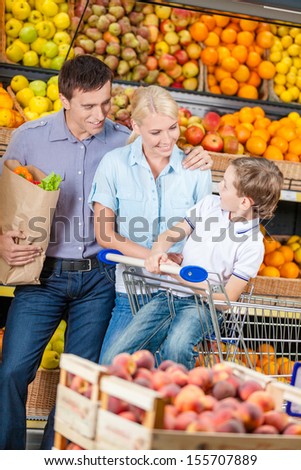 Happy family against shelves of fruits goes shopping. Father keeps a bag with fruits and son sits in the cart