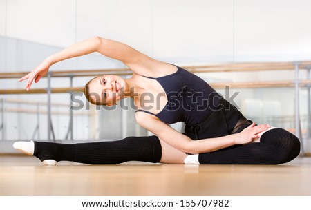 Bending female ballet dancer stretches herself on the floor in the classroom