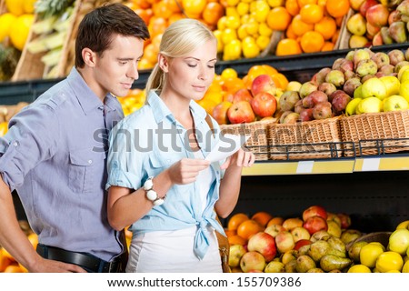 Happy couple with shopping list against the piles of fruits decides what to buy