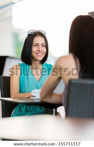 Girl wearing blue dress and sunglasses sits at the table of the cafeteria and talks with friend