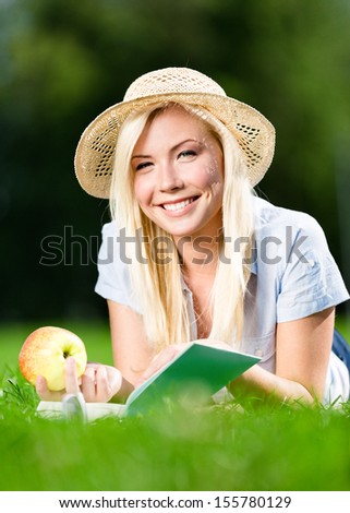 Wearing straw hat girl with healthy apple reads interesting book lying on the green grass in the park