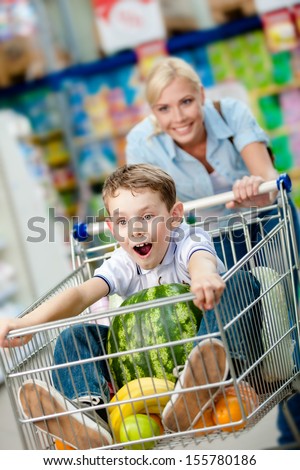 Little boy sits in the shopping trolley with watermelon and other products while mother drives it