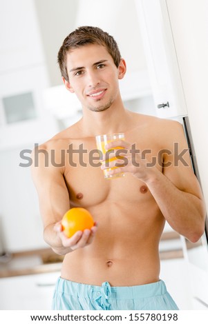 Half-naked man with glass of juice and orange standing near the refrigerator at the kitchen