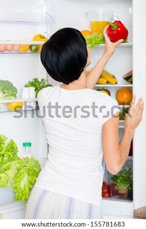 Woman takes red pepper from the opened refrigerator full of vegetables and fruit. Concept of healthy and dieting food