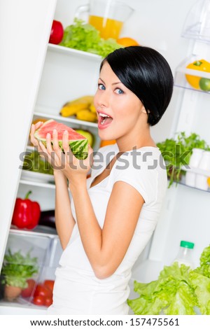 Woman eats watermelon near the opened refrigerator full of vegetables and fruit. Concept of healthy and dieting food