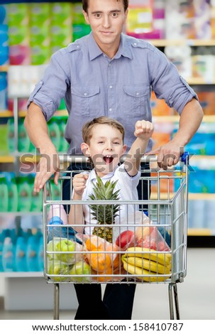 Little Boy With Fists Up Suiting In Shopping Trolley With Food, Father Drives The Cart