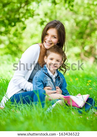 Mom and son with book sitting on green grass in park. Concept of happy family relations and carefree leisure time