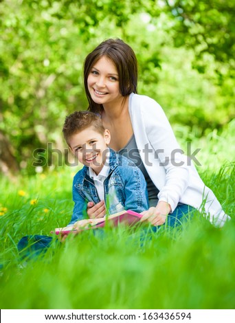 Mother and son with book sitting on green grass in green park. Concept of happy family relations and carefree leisure time