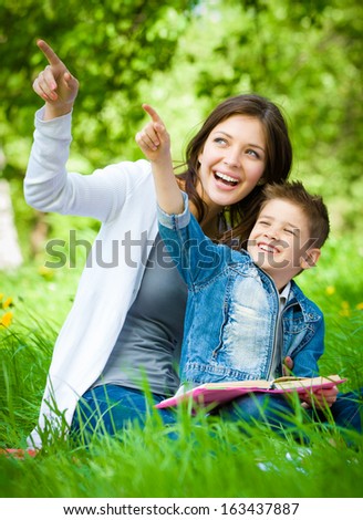 Mother and son with book sitting on green grass pointing hand gesture in park. Concept of happy family relations and carefree leisure time