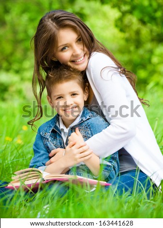 Mum And Son With Book Sitting On Green Grass In Green Park. Concept Of Happy Family Relations And Carefree Leisure Time