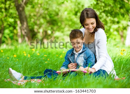 Mother and son with book sitting on green grass in park. Concept of happy family relations and carefree leisure time