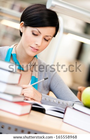 Woman with green apple surrounded with piles of books reads sitting at the table at the library. Academic achievement
