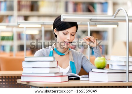 Woman with green apple surrounded with piles of books reads sitting at the desk at the library. Academic achievement