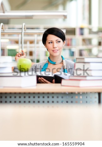 Female student with green ripe apple studies sitting at the desk at the reading hall of the library. Education process