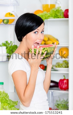 Woman eats watermelon near the opened fridge full of vegetables and fruit. Concept of healthy and dieting food