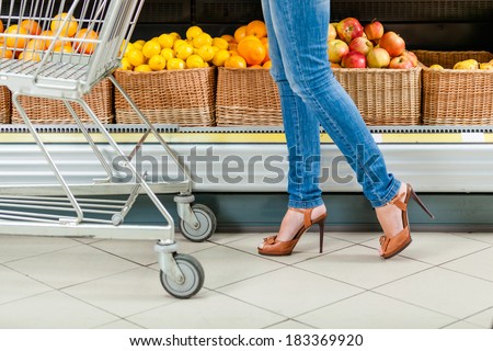 Legs of the female customer with cart against the shelves of fruits in the shop