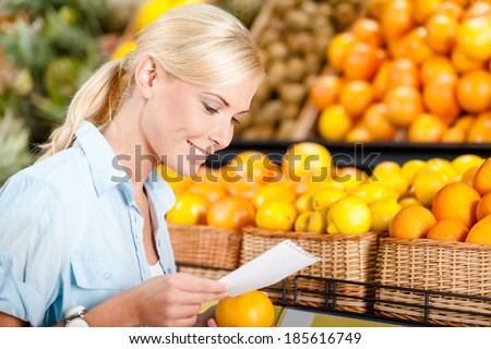 Girl looks through shopping list near the stack of fruits lying in the braided baskets in the store