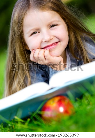 Girl reads interesting book lying on the green grass in the summer park. Red apple is under the book