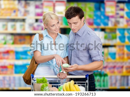 Happy couple discussing the shopping list and chosen products standing near the cart full of food