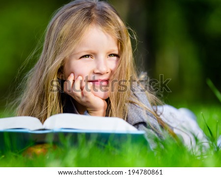 Pretty little girl reads interesting book lying on the green grass in the summer park