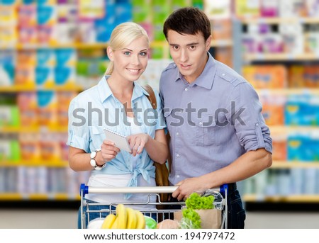 Happy couple discussing the shopping list and chosen products standing near the shopping trolley full of food