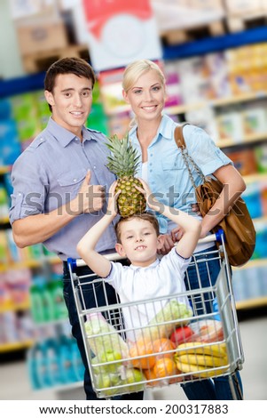 Family drives shopping trolley with food and son sitting there with pine apple on head. Concept of fresh and healthy food and consumerism