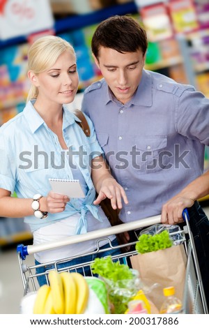 Couple discussing the shopping list and chosen products standing near the shopping trolley full of food