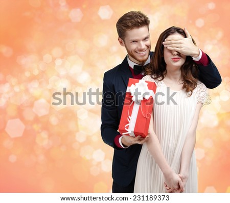 Giving a christmas present man covers eyes of his pretty girlfriend, gold light background