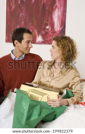 Couple sitting on sofa, unwrapping Christmas present, face to face