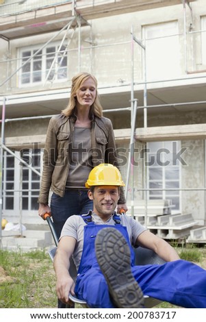 Woman and construction worker at site construction worker sitting in wheelbarrow woman pushing it