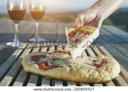 Italy, Tuscany, Magliano, hand holding pizza slice with wine glass in background