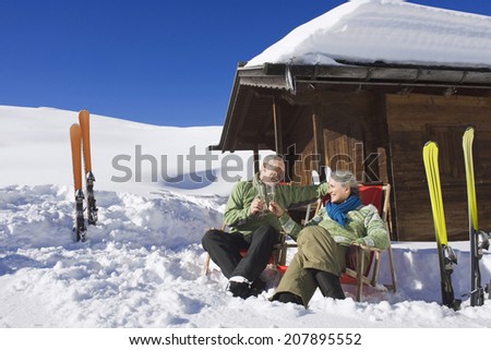 Italy, South Tyrol, Seiseralm, Senior couple sitting in front of log cabin, holding champagne glasses