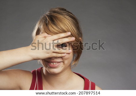 Teenage girl in red tank top looking through fingers smiling against gray background