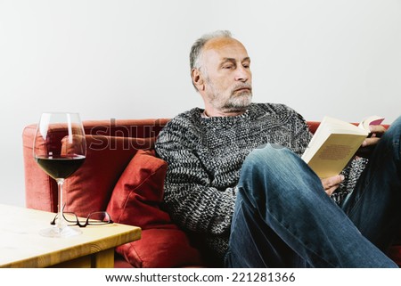 Senior man sitting on couch reading book wine glass with red wine standing on table next to him
