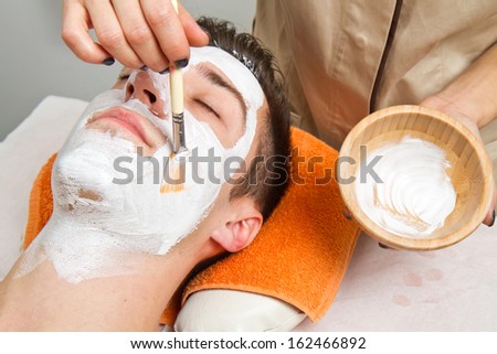 Therapist applying a face mask to a beautiful young man in a spa using a cosmetics brush