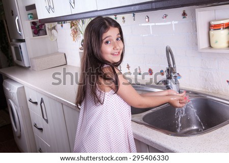 One beautiful middle eastern little girl with pink dress and long dark brown hair and eyes on white kitchen,helping parents to wash dishes and drinking water and smiling\
looking at camera\
studio shot.