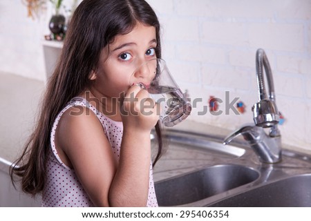 One beautiful middle eastern little girl with pink dress and long dark brown hair and eyes on white kitchen,helping parents to wash dishes and drinking water and smiling\
looking at camera\
studio shot.