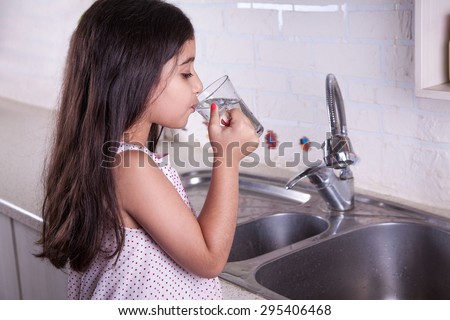 One beautiful middle eastern little girl with pink dress and long dark brown hair and eyes on white kitchen, helping parents to wash dishes and drinking water and smiling. \
studio shot.
