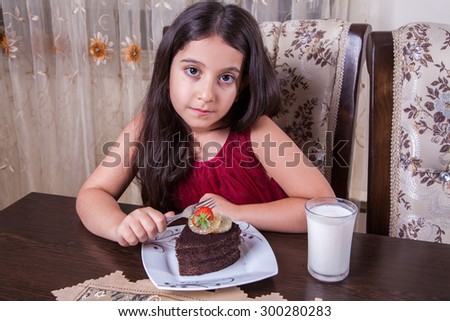 Small beautiful middle eastern girl with chocolate cake with pineapple, strawberry, milk, red dress, dark eyes, long hair, drinking, eating at home, sitting, smiling. looking at camera. studio shot.