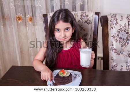 Small beautiful middle eastern girl with chocolate cake with pineapple, strawberry, milk, red dress, dark eyes, long hair, drinking, eating at home, sitting, smiling. looking at camera. studio shot.