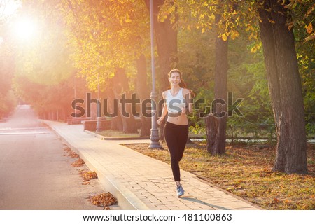 young beautiful happy and sexy runner girl with nice body in the autumnal colored park. with sun on backlight is looking at camera with happiness and smile with white sport suit.