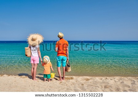 Family on beach wearing straw sun hat. Young couple with three year old boy, holding beach bag and bucket toy. Clear sky. Summer family vacation. Sithonia, Greece.
