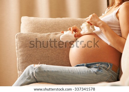 Pregnant woman holding small baby shoes relaxing at home on couch