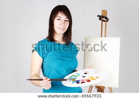 young girl in blue shirt standing near easel, holding brush and palette, smiling