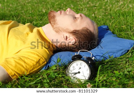 man with beard in yellow shirt sleeping on summer meadow near clock, lying on back, rest concept