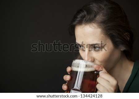 Beautiful young woman with brown hair and brown eyes drinking a mug of beer with a black background.