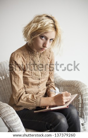 Portrait of a blonde woman with blue eyes sitting in her wicker chair writing in a journal.