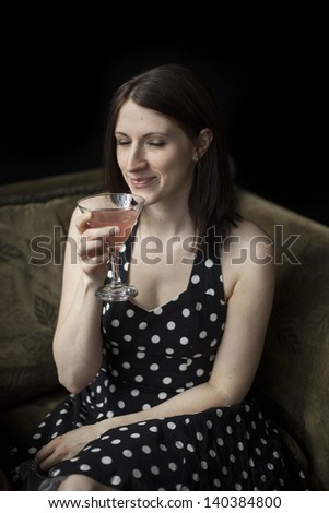 Beautiful young woman with brown hair and eyes in polka dot dress on black background. She is drinking a pink martini.